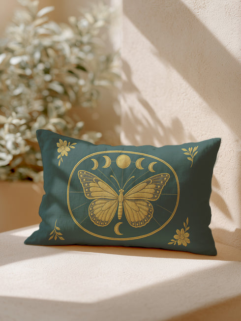 A rectangular lumbar pillow cover, measuring 20 by 12 inches, is displayed against a neutral background. The pillow cover is made of a deep teal green fabric with a subtle, linen-like texture. In the center, a large, intricately detailed butterfly with spread wings is printed in a shimmering gold color. The butterfly is framed by a golden oval which also contains a series of gold moon phase symbols, showing the full lunar cycle. The four corners of the pillow is adorned with gold floral design.